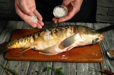 Eller ahşap kesme tahtasındaki taze yakalanmış balığın üzerinde deniz tuzu tutar. Mutfak koyu tonları ile taşralıdır. Bitkiler ve baharatlar balıkların etrafını sarmış, baharata hazır..