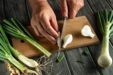 Eller, ahşap kesme tahtasındaki taze soğanları ustalıkla dilimler. Kurulun etrafında taze yemek malzemelerini vurgulayan taze soğanlar var..