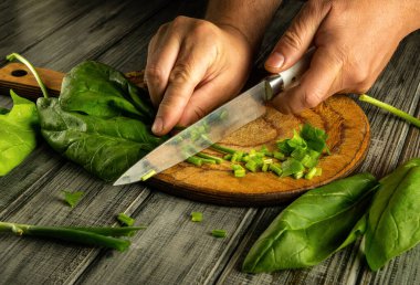 Eller ahşap bir tahtada taze ıspanağı ustalıkla doğruyor. Parlak bir mutfakta sağlıklı bir yemek için malzemeleri hazırlıyor. Odak noktamız bıçak ve canlı yeşillikler..