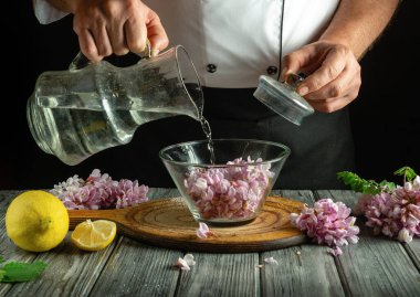 Bir şef, ahşap bir masada taze limon dilimleriyle çevrili leylak yapraklarıyla dolu cam bir kaseye dikkatlice temiz su döker. Sahne sakin ve odaklanmış..