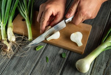 Eller taze taze soğanları tahta bir tahtanın üzerinde doğruyor. Mutfak doğal ışıkla parlıyor, sebzelerin canlı renklerini vurguluyor..