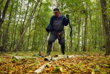 Bir adam yemyeşil bir ormanda metal dedektörüyle gömülü hazineleri arıyor. Güneş ışığı ağaçların arasından süzülerek doğada sakin bir atmosfer yaratır..