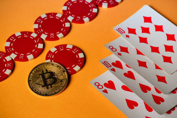Red poker chips and playing cards are spread out on an orange surface. A bitcoin coin sits next to the cards, suggesting an online gaming or betting theme.