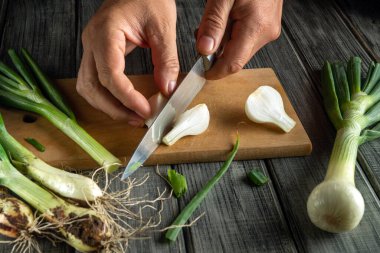 Tahta bir kesme tahtasının üzerinde taze soğan doğrayan eller görülüyor. Mutfak aydınlık, taze sebzeleri ve kesim sürecini ön plana çıkarıyor..
