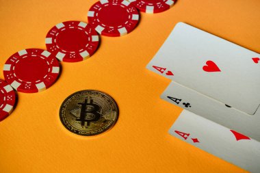 Canlı bir düzenek, kırmızı poker çipleri ve parlak turuncu bir yüzey üzerinde bitcoin ile birlikte iki as gösteren üç oyun kartı gösteriyor, dinamik bir oyun atmosferi yaratıyor..