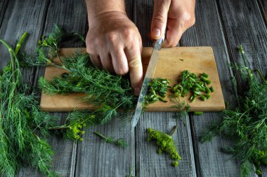 Bir insan ahşap bir kesme tahtasının üzerinde ustalıkla taze otları doğruyor. Tahtanın etrafında çeşitli otlar var, canlı yeşil renkler sergiliyorlar. Doğal ışık mutfağı aydınlatır..