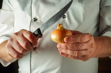 Bir şef bir elinde küçük bir soğan tutarken diğer elinde keskin bir bıçak tutuyor. Sahne iyi aydınlatılmış bir mutfakta geçiyor, taze malzemeler sergileniyor..