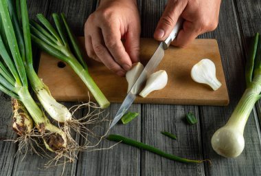 Taze sebzelerle çevrili ahşap bir tahtanın üzerindeki yeşil soğanları ustalıkla dilimle dilimle. Sahne, sıcak bir mutfakta yemek hazırlama anını yakalıyor..