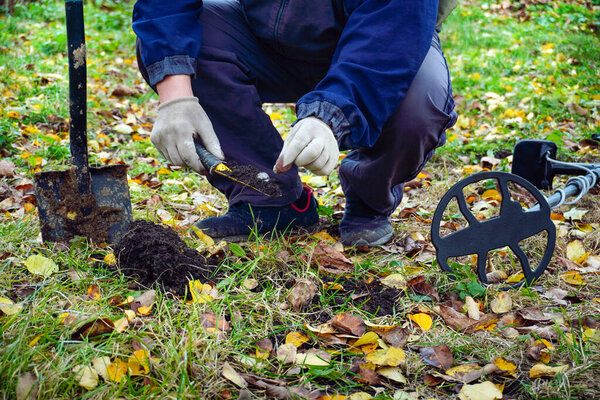 Человек преклоняет колени в осеннем парке, используя металлодетектор и небольшую лопату для поиска спрятанных сокровищ. Красочные листья покрывают землю, создавая яркую сцену.