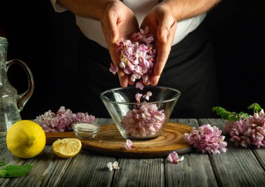 Elleri cam bir kasenin üzerinde taze mor çiçekleri tutar ve onları bir tabakta kullanılmak üzere hazırlarlar. Limon, tuz ve otlar kasenin etrafını sararak lezzetli bir yemek pişirme sürecini ön plana çıkarıyor..