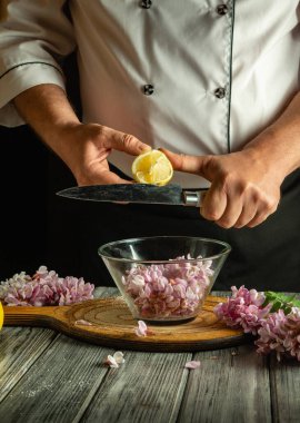 Bir şef ustalıkla limon suyunu narin çiçek yapraklarıyla dolu bir cam kaseye sıkıyor. Taze malzemeler sıcak mutfakta canlı, aromatik bir atmosfer yaratıyor..