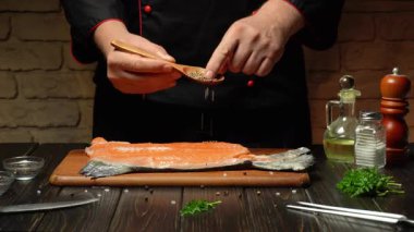 Sıcak bir mutfakta, bir şef somon filetosunu dikkatlice baharatlarla doldurur ve ahşap kaşık kullanır. Zeytinyağı ve bitkiler yakında, kullanıma hazır..