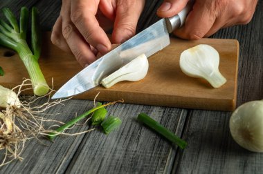 Taze soğanları ahşap bir tahtanın üzerinde ustalıkla doğrayan eller taze ürünleri ve basit mutfak aletlerini sergiliyor. Bu sahnede lezzetli bir yemek hazırlığı vurgulanıyor..