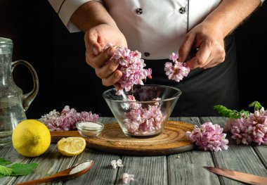 Bir şef cam bir kaseye dikkatlice yenilebilir çiçek yaprakları ekler, limon ve ahşap bir kesme tahtasındaki şifalı bitkiler gibi taze malzemelerle kaplıdır..