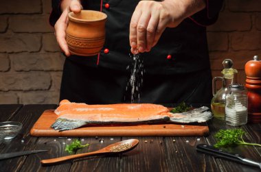 Bir şef, taze bir somon balığını tuzlu bir mutfakta bekleterek mevsim geçirir. Malzemeler ve aletler ahşap bir masaya güzelce dizilir ve davetkar bir atmosfer yaratır..