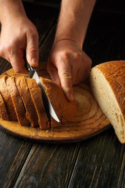 Eller, ahşap bir kesme tahtasıyla taze pişmiş bir ekmeği dikkatlice dilimlerken görülüyor. Sahne, sıcak bir mutfakta ev yemeğinin sıcaklığını ve rahatlığını yakalıyor..
