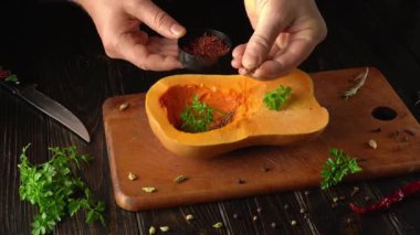 Eller dikkatle baharatları ahşap bir kesme tahtasının üzerinde yarım yamalak bir kabağa serpiyor. Yakınlarda taze otlar ve bıçak var. Bu da mutfağın sıcak atmosferinde yemek hazırlama sahnesi olduğunu gösteriyor..