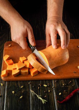 Bir insan tahta bir kesme tahtasıyla kabak ya da balkabağını küp şeklinde kesiyor. Çeşitli baharatlar etrafa saçılmış, canlı bir mutfak sahnesi yaratıyorlar..