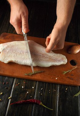 Elleri, taze bitkiler ve baharatlarla çevrili ahşap bir tahtanın üzerinde ustalıkla taze balık filetosu şeklinde kesilmiş. Sıcak mutfak atmosferi yemek pişirme deneyimini arttırır..
