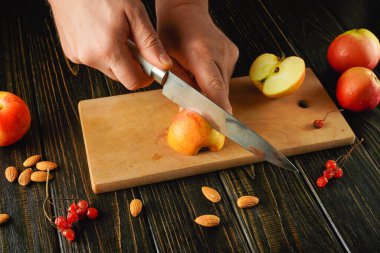 Elleri tahta bir kesme tahtasında kırmızı bir elmayı dilimliyor. Sahne dağınık badem ve kırmızı üzümleri içeriyor. Sıcak bir mutfakta sağlıklı bir aperatif oluşturuyorlar..