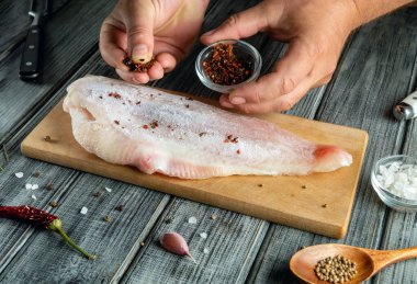 Eller, ahşap bir tahtaya yerleştirilmiş tuzlu balık filetosunun üzerine baharat serpiyor. Kurulumu sarımsak ve kırmızı biber gibi taze malzemeler sarar. Aşçılık sıcak bir mutfak atmosferinde yapılır..