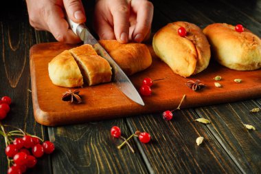 Taze pişmiş zanaatkar ekmeği ahşap bir kesme tahtasıyla dilimleniyor. Ekmek altın kahverengidir ve yanında canlı kirazlar ve aromatik baharatlar ile sıcak bir mutfak sahnesi yaratır..