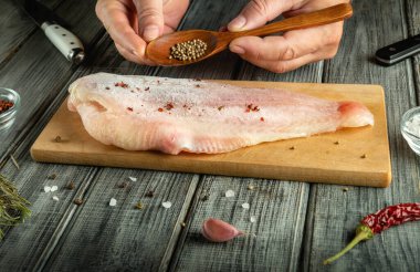 Elleri taze balık filetosunu, mutfaktaki tahta kesme tahtasındaki baharatlarla tatlandırıyor. Yakınlarda yemek pişirmek için çeşitli malzemeler var..