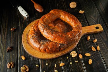 Taze ekmek ya da simit ahşap kesim tahtasının üzerinde haşhaş tohumlarıyla birlikte durur. Baharatlar ve fındıklar tahtaya saçılmış. Yakınlarda da bir bıçak var..