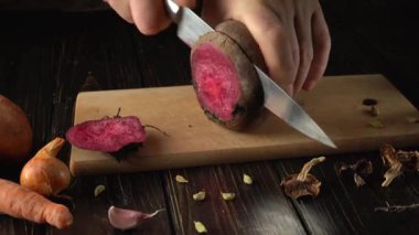 Bir el tahta bir tahtanın üzerinde tatlı bir patatesi keser. Yakınlarda havuç ve sarımsak gibi sebzeler var. Mutfak, pencereden gelen doğal ışıkla aydınlanmış..