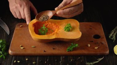 Bir insan içi oyulmuş tereyağlı kabağa tohum eklemek için tahta kaşık kullanır. Taze otlar kabağın içinde duruyor ve bir bıçak da tahta kesme tahtasının yanında..