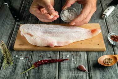 Elleri ahşap kesme tahtasındaki balık filetosuna tuz ekliyor. Balıkların etrafına baharatlar ve bitkiler konur. Ayarlar yemek pişirme aletleriyle bir mutfağı gösteriyor..