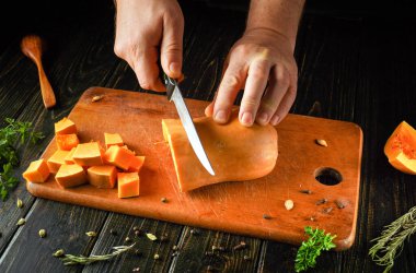 Elleri ustalıkla olgun bir tereyağlı kabağı tahta bir kesme tahtasına kesti. Kabağı çevreleyen doğranmış parçalar sıcak bir mutfakta canlı bir pişirme sürecini vurguluyor..