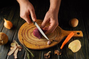 Bir insan, sıcak bir mutfak atmosferinde çeşitli taze sebzeler ve malzemelerle çevrili ahşap bir kesme tahtasının üzerinde ustalıkla kırmızı bir pancar doğruyor..