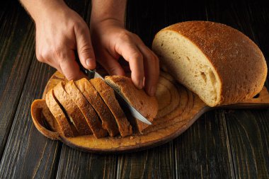 Elleri bir bıçak tutuyor, bir somun taze ekmeği tahta bir kesme tahtasıyla dilimliyor. Sıcak, kırsal mutfak atmosferi bu aktivitenin rahatlatıcı duygusunu arttırıyor..