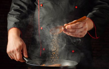 Bir şef sobanın tepsisine baharat serpmek için tahta kaşık kullanır. Akşam vakti mutfak ortamında pişirilen yemeklerden buhar yükselir..