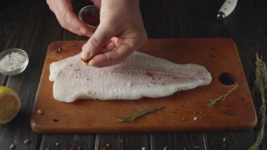 Tahta bir kesme tahtasının üzerine çiğ balık filetosu serpiştirilmiş bir el. Yakınlarda tuz, şifalı otlar ve masada bir limon var. Bıçak da görünür..