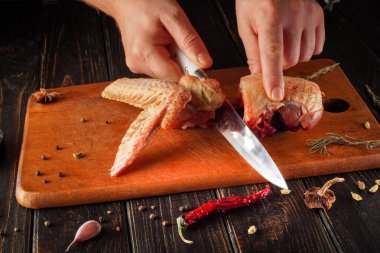 Eller ahşap bir kesme tahtasıyla çiğ tavuk kanadı kesiyor. Tahtanın her tarafına çeşitli otlar ve baharatlar yayılmış. Faaliyet bir mutfak ortamında gerçekleşiyor..