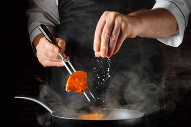 Bir aşçı bir parça yemeği çatalla kızartma tavasının üzerinde tutar. Etrafına buhar yükselirken, tabağa tuz serpiyor. Sahne yemek sırasında kalabalık bir restoran mutfağında geçiyor..