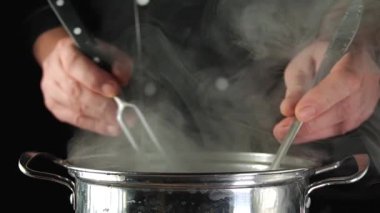 Sobanın üzerinde çatal ve kepçe tutan iki el, buhar yükseliyor. Bir kişi yemek pişirirken mutfakta yemek hazırlıyor..