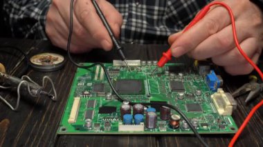 İki el, kırmızı ve siyah sondalı çoklu metre kullanarak devre kartındaki bileşenleri ölçüyor. Araçlar ve diğer küçük parçalar masada görünür..