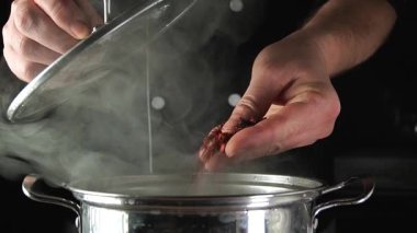 Eller sobadaki tencereye kurutulmuş malzemeler ekliyor. Demlikten buhar yükseliyor, mutfak ortamında pişirme sürecini gösteriyor..