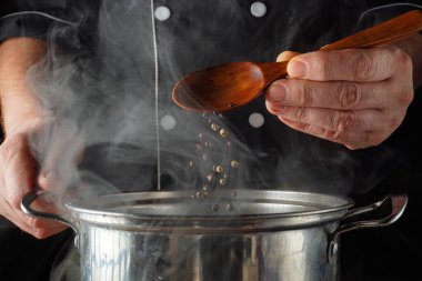 Bir şef, mutfak ortamında sobanın üstündeki buharlı tencereye baharat ya da kuru biber döker. Şef yemek yapmaya odaklanmış..