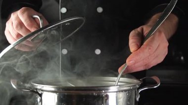 Bir aşçı mutfakta duruyor, ocağın üstündeki bir tencerede bir kepçeyle kaynar su karıştırıyor. Buhar yükselir, boşluğu sıcaklıkla ve yiyecek kokusuyla doldurur. Metal kaplar mutfaktan gelen ışığı yansıtıyor..