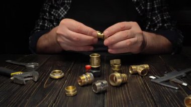Birkaç yedek parça masada yatarken bir tesisatçının elleri pirinçleri tamir ediyor. İngiliz anahtarı ve kalibre gibi araçlar yakınlardadır. Bu sahne bir atölyedeki tesisat çalışmalarını gösteriyor..