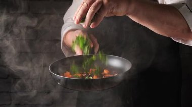 Bir şef mutfakta yemek pişirirken renkli sebzelerle tavaya taze otlar ya da maydanoz serper. Şef tabağa odaklanırken tavadan buhar yükseliyor..