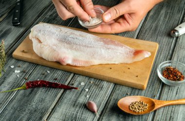 Tahta kesme tahtasının üzerinde duran balık filetosuna tuz ekleniyor. Yakınlarda çeşitli baharatlar, otlar ve mutfak tezgahında bir bıçak var..