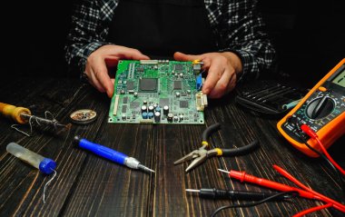 Tahta bir masada oturan bir insan devre kartında çalışır. Levye ve çoklu metre gibi aletler yakında. Dekor loş, akşam vaktini gösteriyor..