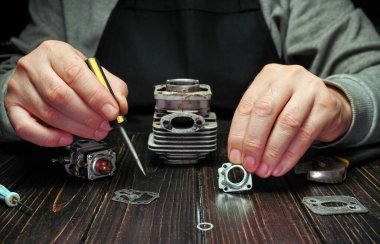 Tahta bir masadaki motor parçaları üzerinde çalışmak için bir tornavida kullanıyor. Çevrelerinde çeşitli araçlar ve mekanik bileşenler düzenlenir. Sahne akşam çekiliyor..