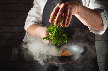 Şef tavaya taze maydanoz ekleyerek yemek hazırlar. Sahne, mutfak ortamında tavadan yükselen dumanı gösteriyor. Yemek pişirme sürecine ve malzemelere odaklanıyoruz..