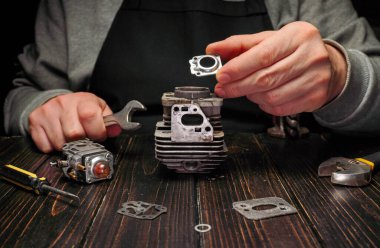 Bir insan ahşap bir masaya yerleştirilmiş motor parçaları üzerinde çalışır. Cihazlar yakınlarda. Çalışmada, akşam boyunca bileşenlerin montajı ve onarımı üzerinde duruluyor..